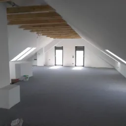 Poddasze z szarą wykładziną dywanową, widoczne drewniane elementy konstrukcyjne dachu, okna dachowe i dwa okna pionowe z żaluzjami, ściany pomalowane na biało, w tle wiadro z farbą.