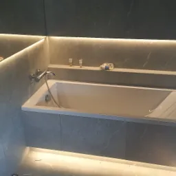 Nowoczesna łazienka z wanną obudowaną szarymi płytami imitującymi marmur, podświetlona taśmami LED, z chromowaną armaturą i półką na kosmetyki.