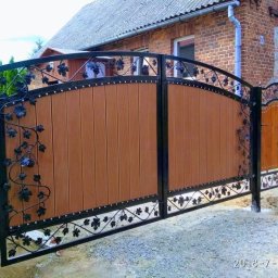 PPHU LAND-MET Dariusz Landmesser - Dwuskrzydłowa brama wjazdowa z drewnianym wypełnieniem i metaloplastyką z motywem winogron, na tle ceglanego domu. Widoczny fragment słupka z jasnego kamienia.