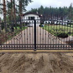 PPHU LAND-MET Dariusz Landmesser - Kuta, dwuskrzydłowa brama wjazdowa z dekoracyjnymi elementami, zamontowana na ceglanych słupkach. W tle dom i drzewa. Widoczne ślady opon na ziemi.
