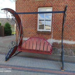 PPHU LAND-MET Dariusz Landmesser - Metalowa huśtawka ogrodowa z drewnianym siedziskiem, zdobiona ornamentami, na tle ceglanego budynku i kostki brukowej. Widoczne detale wykonania i otoczenie.