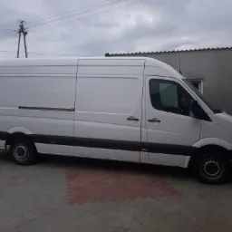 Biały samochód dostawczy zaparkowany na kostce brukowej, prawdopodobnie transportujący sprzęt spawalniczy lub materiały potrzebne do spawania.