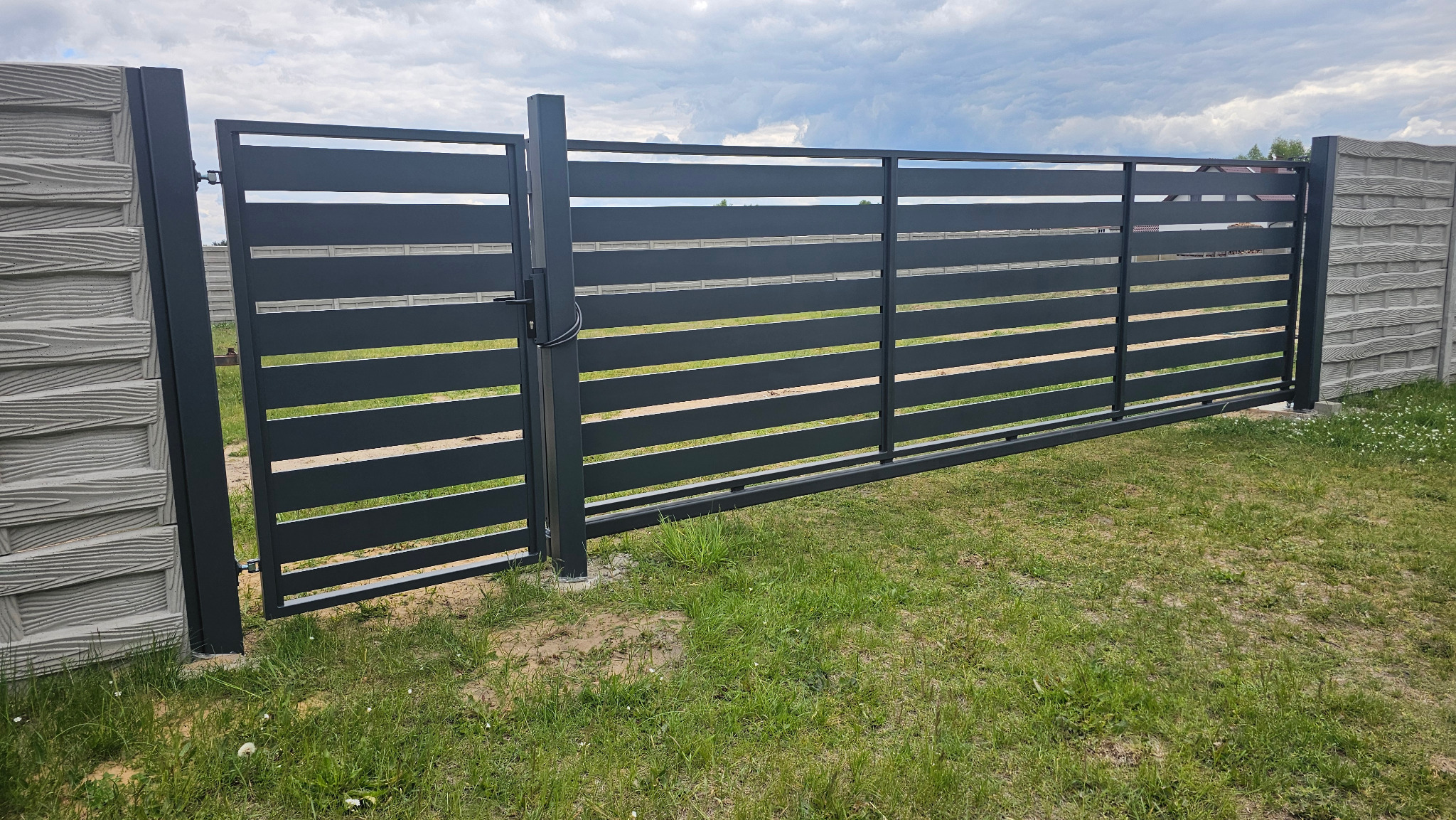 Nowoczesna, przesuwna brama posesyjna z poziomymi panelami w kolorze antracytowym, zamontowana między betonowymi słupkami imitującymi drewno, na tle zielonego trawnika.