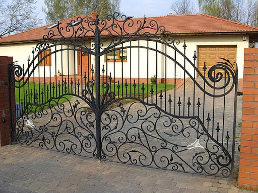 Kuta, ozdobna brama wjazdowa z metalu, na tle domu z czerwoną dachówką. Precyzyjne zdobienia i solidna konstrukcja. Widok z zewnątrz.