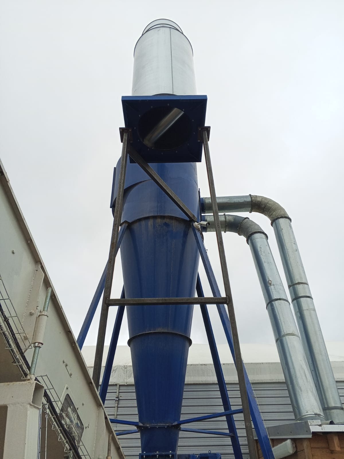 Niebieski cyklon przemysłowy z metalowymi rurami, wsparty na stalowej konstrukcji, sfotografowany z dołu na tle pochmurnego nieba. Widoczne elementy spawane.