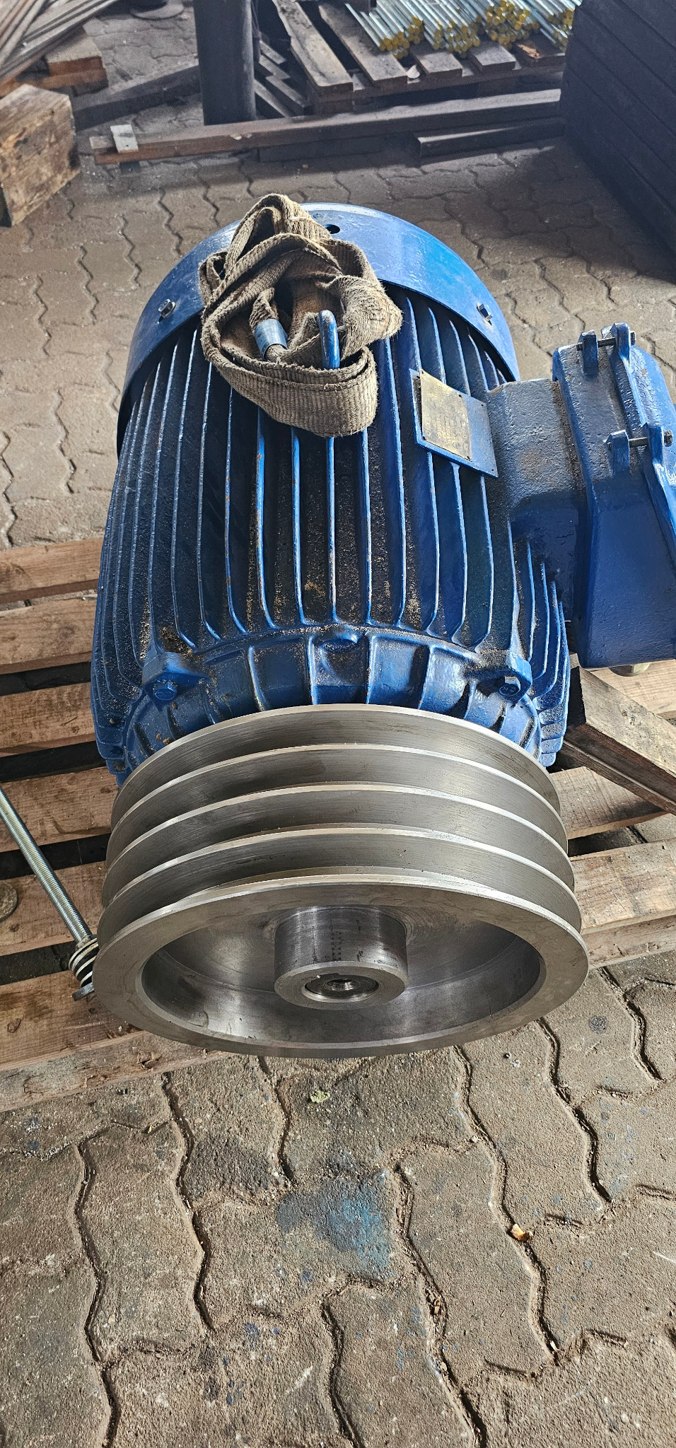 Niebieski silnik elektryczny z kołem pasowym na drewnianej palecie, widoczne ślady użytkowania i szmatka na górze. Detal z warsztatu.