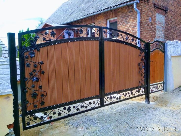 Dwuskrzydłowa brama wjazdowa z drewnianym wypełnieniem i metaloplastyką z motywem winogron, na tle ceglanego domu. Widoczny fragment słupka z jasnego kamienia.