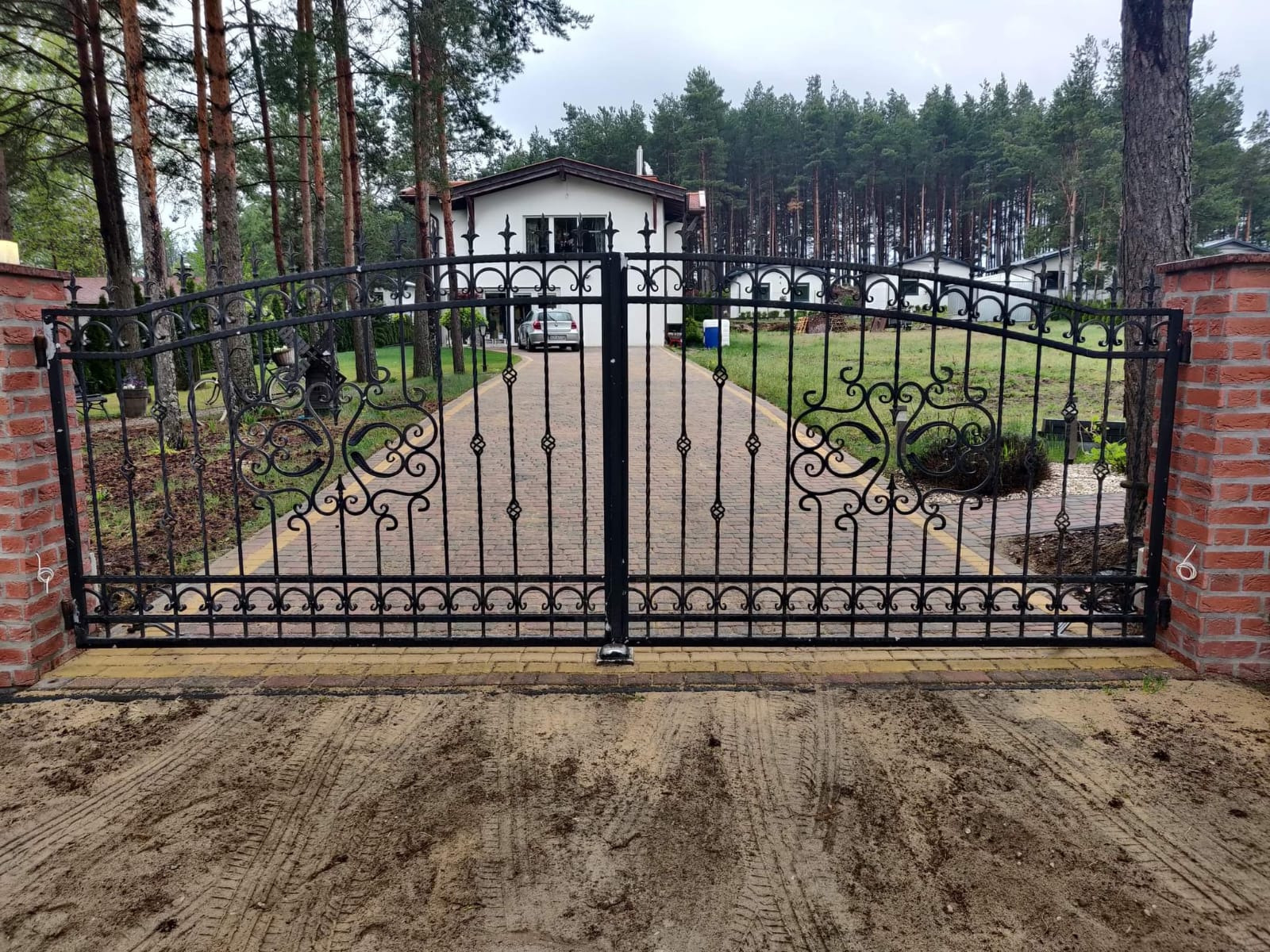 Kuta, dwuskrzydłowa brama wjazdowa z dekoracyjnymi elementami, zamontowana na ceglanych słupkach. W tle dom i drzewa. Widoczne ślady opon na ziemi.