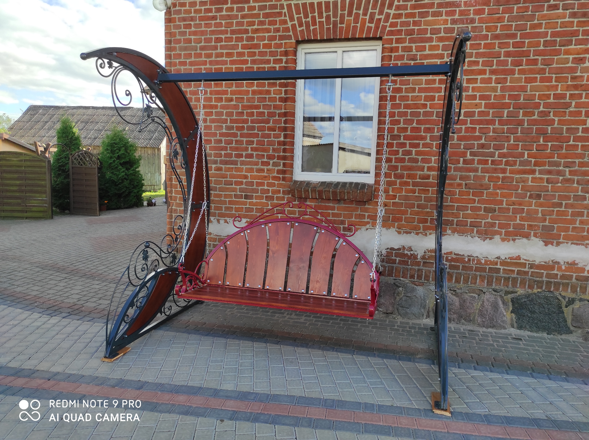 Metalowa huśtawka ogrodowa z drewnianym siedziskiem, zdobiona ornamentami, na tle ceglanego budynku i kostki brukowej. Widoczne detale wykonania i otoczenie.