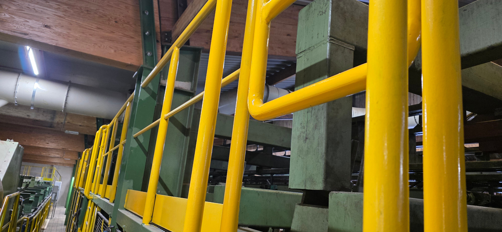 Żółte barierki ochronne na konstrukcji stalowej w hali produkcyjnej. Widoczne elementy spawane i łączenia śrubowe. Oświetlenie jarzeniowe.