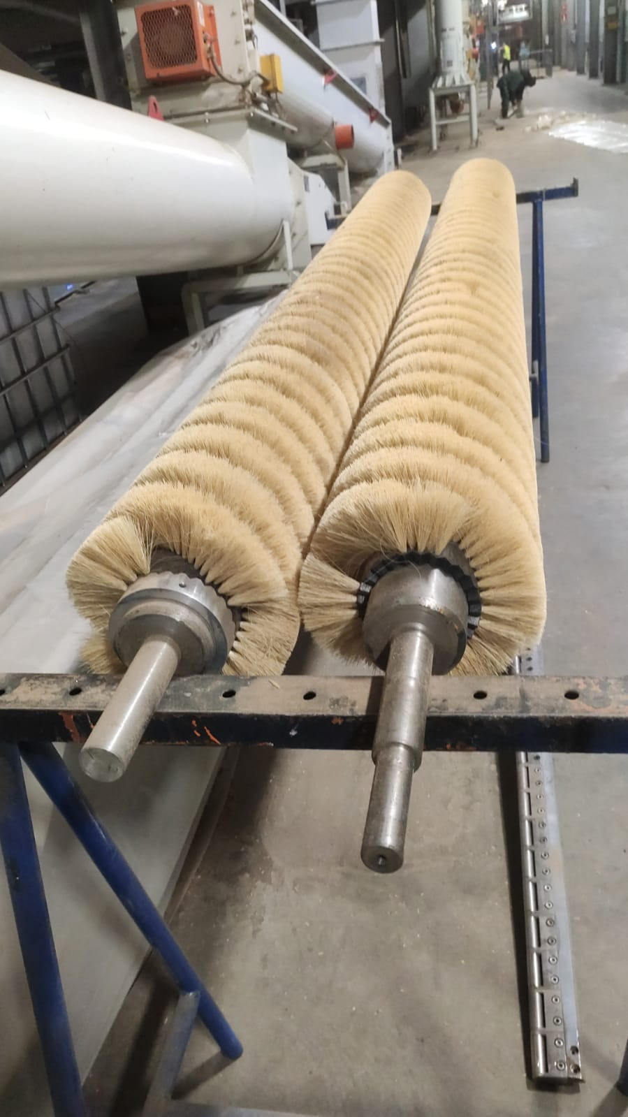 Dwa duże, cylindryczne wałki z włosiem na metalowych osiach, umieszczone na niebieskim stojaku w hali przemysłowej. Widoczne elementy maszyn i pracownik w tle.