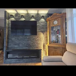 Ściana z szarej okładziny kamiennej z wbudowanym czarnym kominkiem elektrycznym i wiszącym telewizorem, oświetlona czterema reflektorami punktowymi; obok stoi drewniany kredens z zegarem...