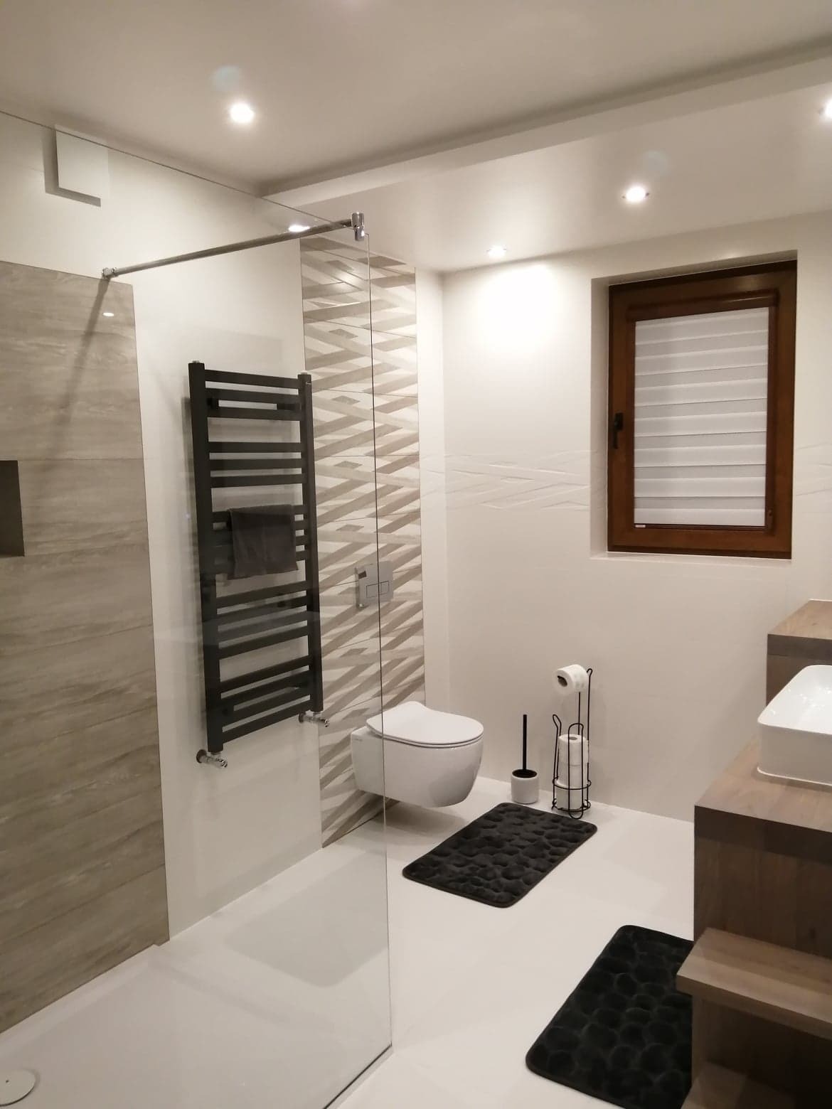 Nowoczesna łazienka z kabiną prysznicową typu walk-in, podwieszaną toaletą, grzejnikiem drabinkowym na ręczniki, drewnianą szafką pod umywalką i oknem z żaluzjami.