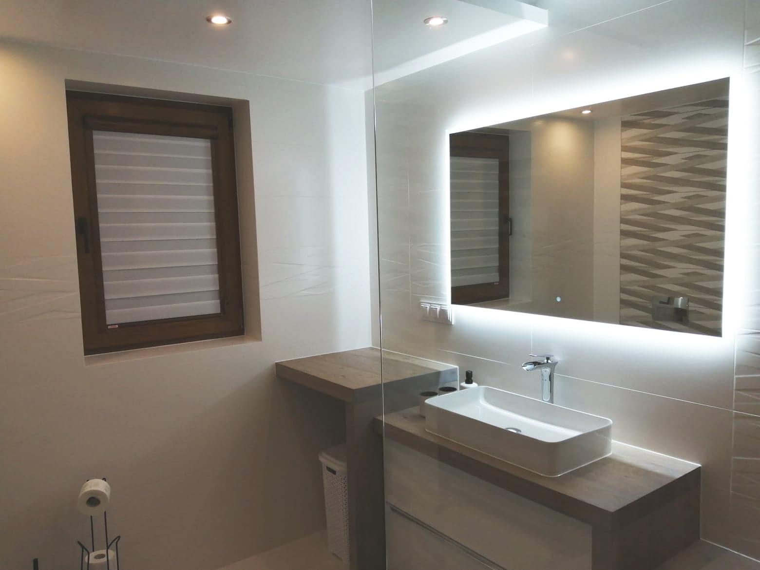 Nowoczesna łazienka z podświetlanym lustrem, prostokątną umywalką nablatową, drewnianym blatem i oknem z żaluzjami poziomymi.