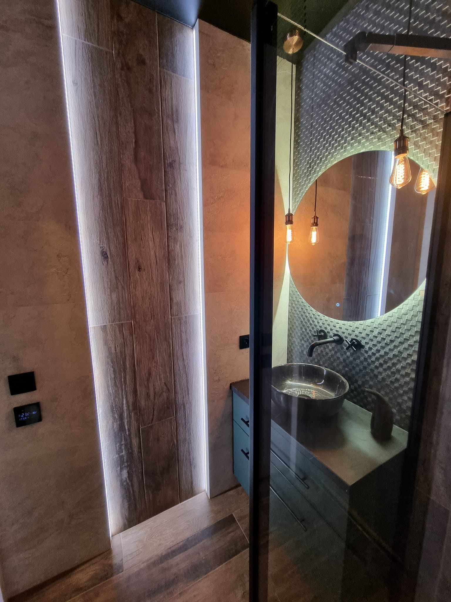 Nowoczesna łazienka z drewnopodobnymi płytkami podświetlonymi LED, okrągłym lustrem z dekoracyjną mozaiką w tle oraz wiszącymi żarówkami Edisona.