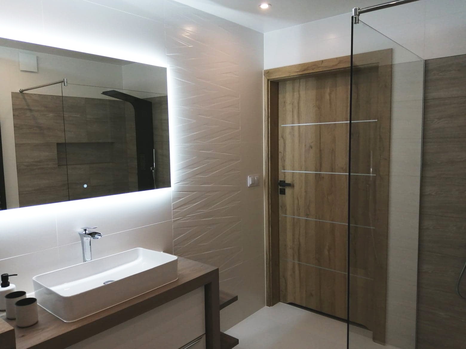 Nowoczesna łazienka z podświetlanym lustrem, prostokątną umywalką nablatową, drewnopodobnymi drzwiami z poziomymi wstawkami i kabiną prysznicową ze szklaną ścianką.