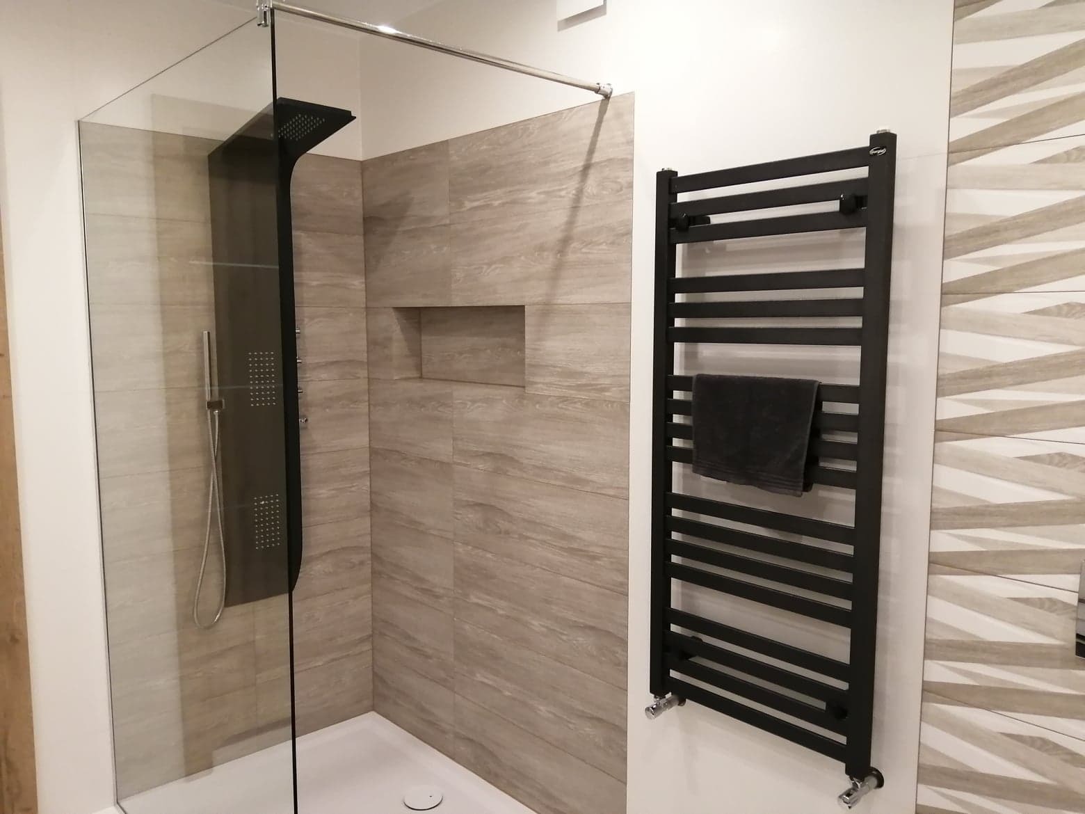 Nowoczesna łazienka z kabiną prysznicową typu walk-in, wykończona płytkami imitującymi drewno i wyposażona w czarny grzejnik drabinkowy z zawieszonym szarym ręcznikiem.