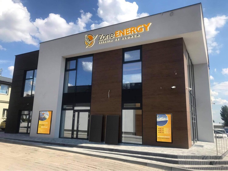 Dwukondygnacyjny budynek firmy Zone Energy z dużym logo na elewacji, reklamami na ścianach i panelami słonecznymi ustawionymi przed wejściem.