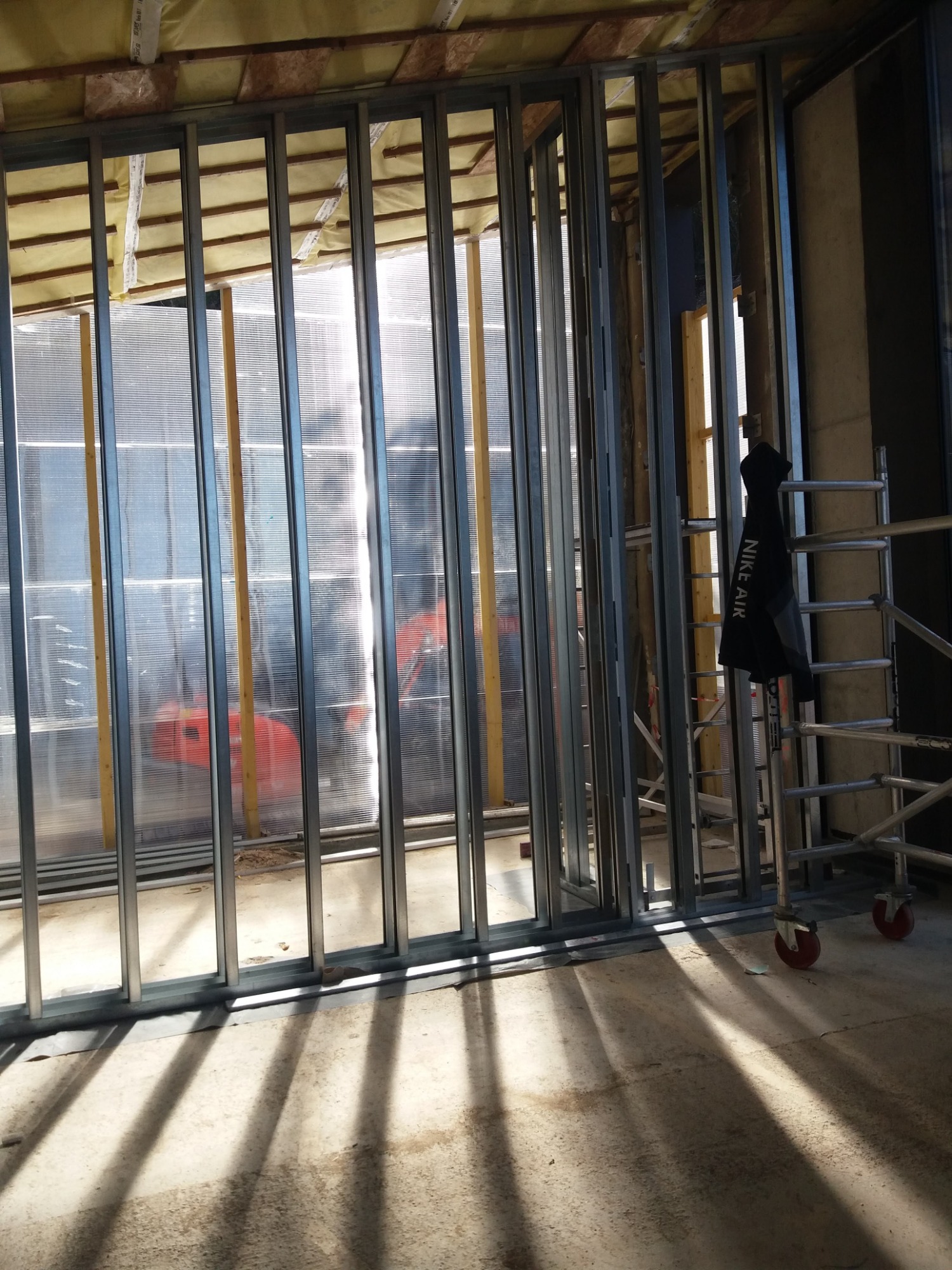 Metalowa konstrukcja szkieletowa ściany z widoczną izolacją i folią, częściowo zasłonięta kurtką na rusztowaniu, w tle czerwony element wyposażenia.