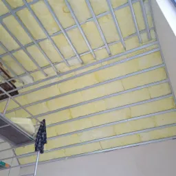 Poddasze w trakcie ocieplania: widoczna wełna mineralna w metalowej konstrukcji nośnej, fragment rusztowania z bluzą w kratę.