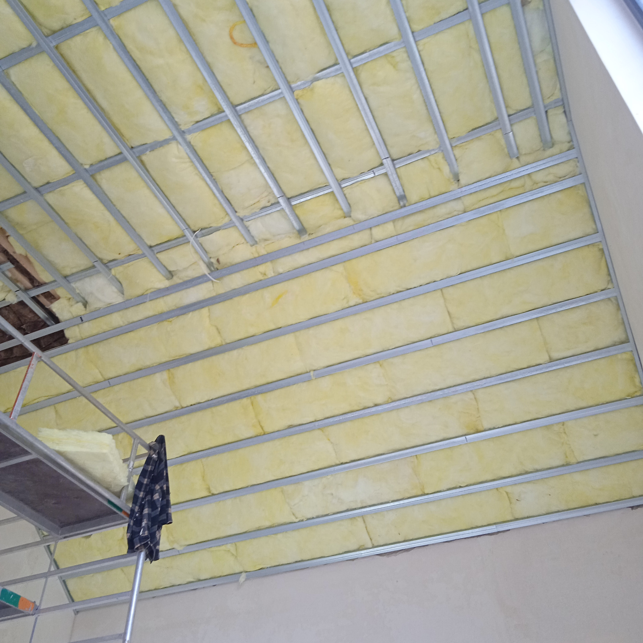 Poddasze w trakcie ocieplania: widoczna wełna mineralna w metalowej konstrukcji nośnej, fragment rusztowania z bluzą w kratę.