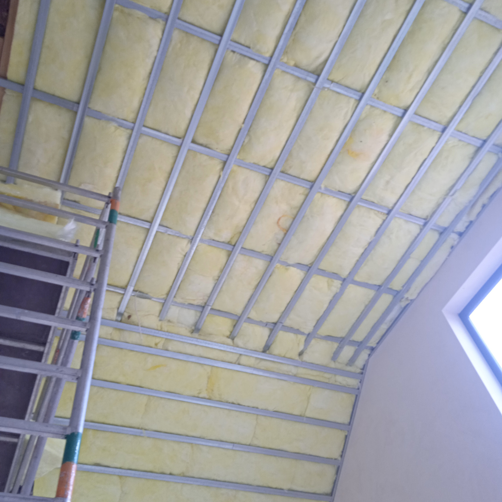 Żółta wełna mineralna w metalowym stelażu sufitowym na poddaszu, widoczne fragmenty drabiny malarskiej i okna.