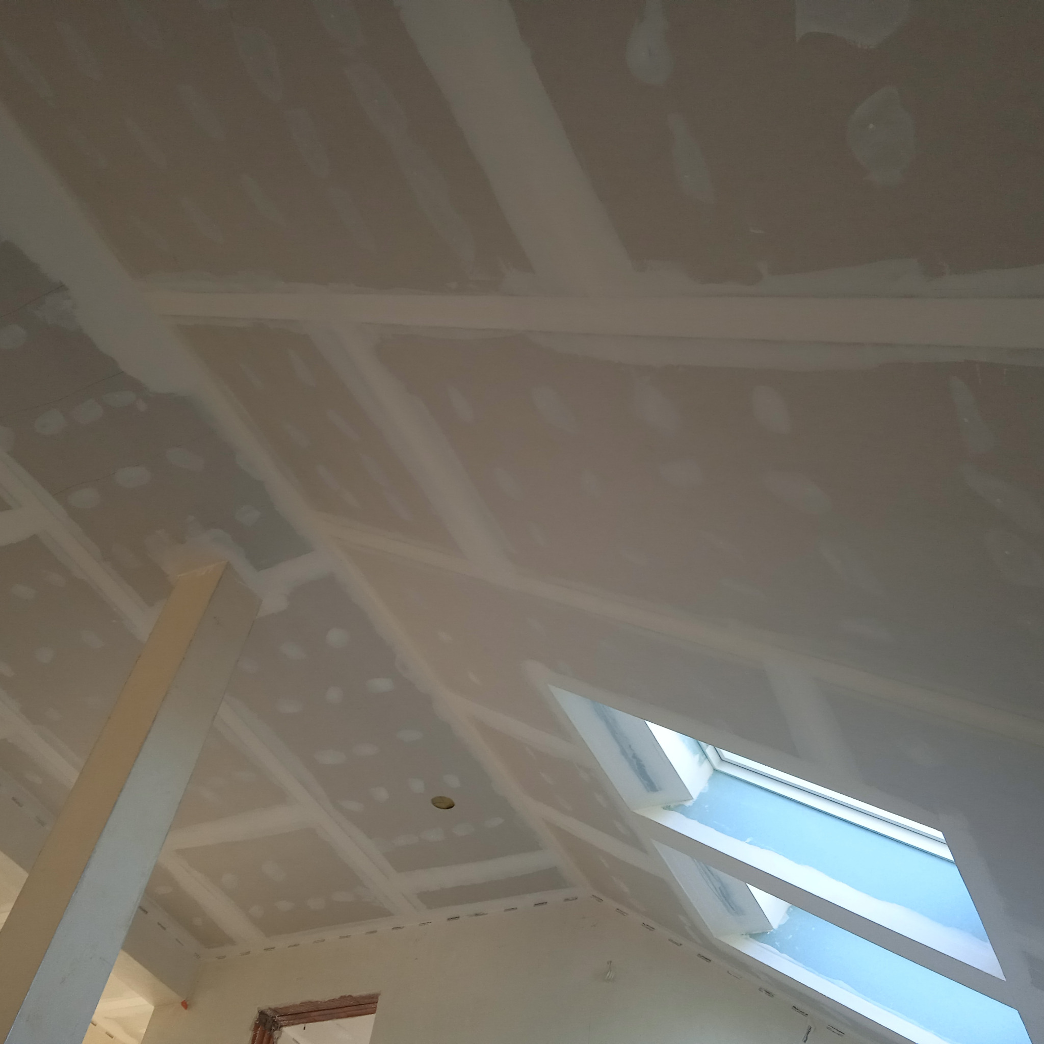 Poddasze z zamontowanymi płytami kartonowo-gipsowymi, widoczne łączenia szpachlowane, okno dachowe w skosie dachu, słup podtrzymujący konstrukcję.