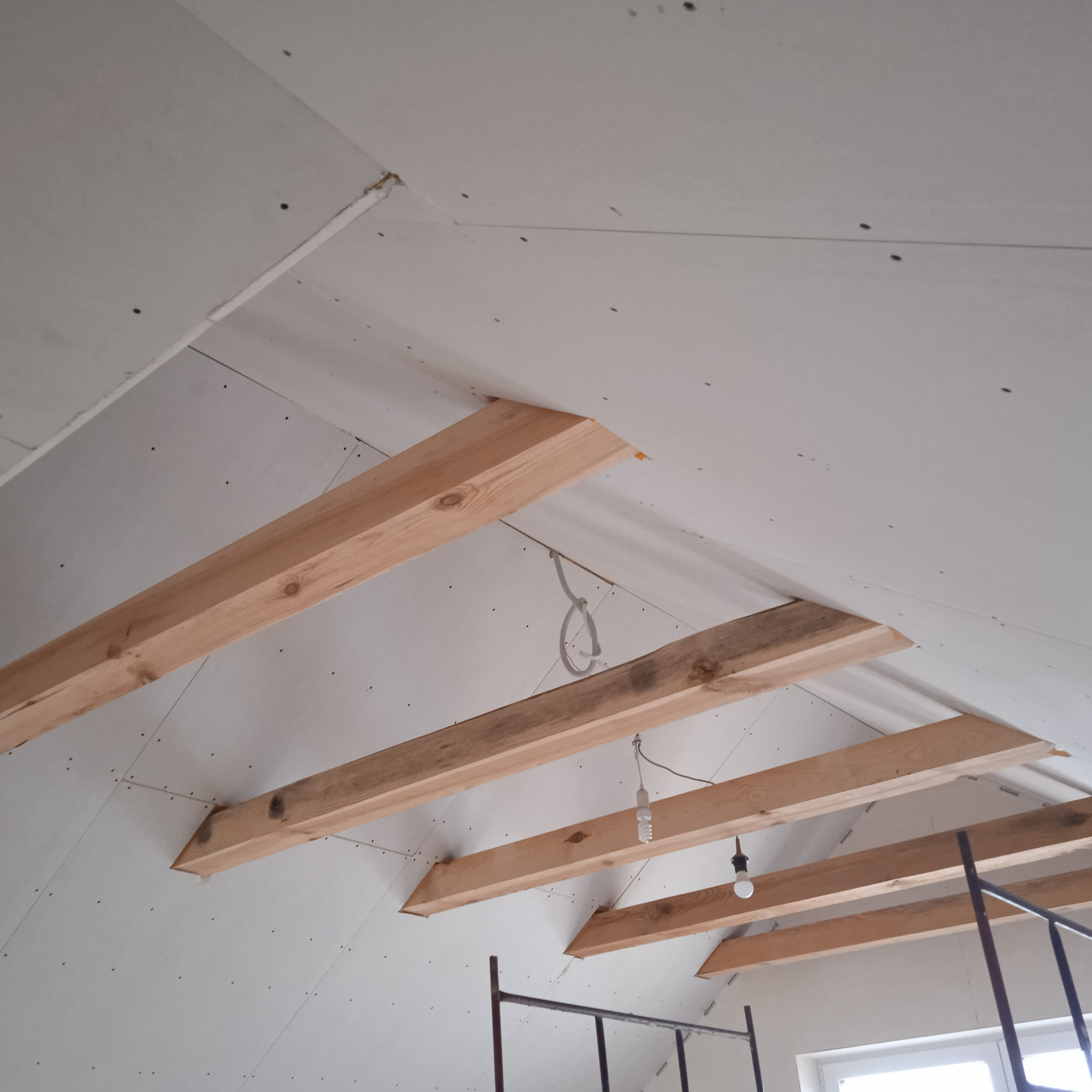 Poddasze w trakcie wykańczania płytami kartonowo-gipsowymi, widoczne drewniane belki konstrukcyjne i częściowo zamontowane płyty, z wiszącą żarówką na kablu i fragmentem rusztowania w dolnej części...
