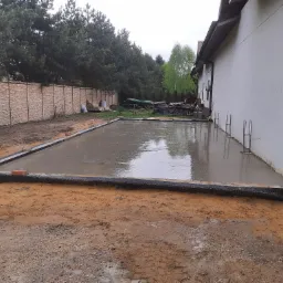 Wylany fundament pod budynek jednorodzinny, widoczne zbrojenia i izolacja przeciwwilgociowa, w tle ogrodzenie z kamienia łupanego i drzewa.