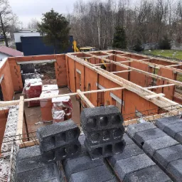 Wznoszenie ścian z czerwonej cegły ceramicznej Porotherm, widoczne zbrojenie i bloczki betonowe na górze, w tle żółta koparka na placu budowy.