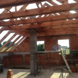 Wnętrze domu w trakcie budowy: widoczne ściany z czerwonej cegły, drewniana konstrukcja dachu, betonowy filar i rusztowanie z niebieskim pojemnikiem na górze.