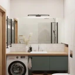 Nowoczesna łazienka z pralką w zabudowie pod drewnianym blatem, zielone szafki, biała umywalka, wisząca toaleta i kosze wiklinowe, odbicie kabiny prysznicowej w lustrze.