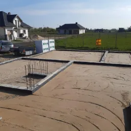 Przygotowany teren pod wylewkę fundamentów domu jednorodzinnego, widoczne zbrojenia i bloczki fundamentowe, w tle zabudowa jednorodzinna i błękitne niebo.