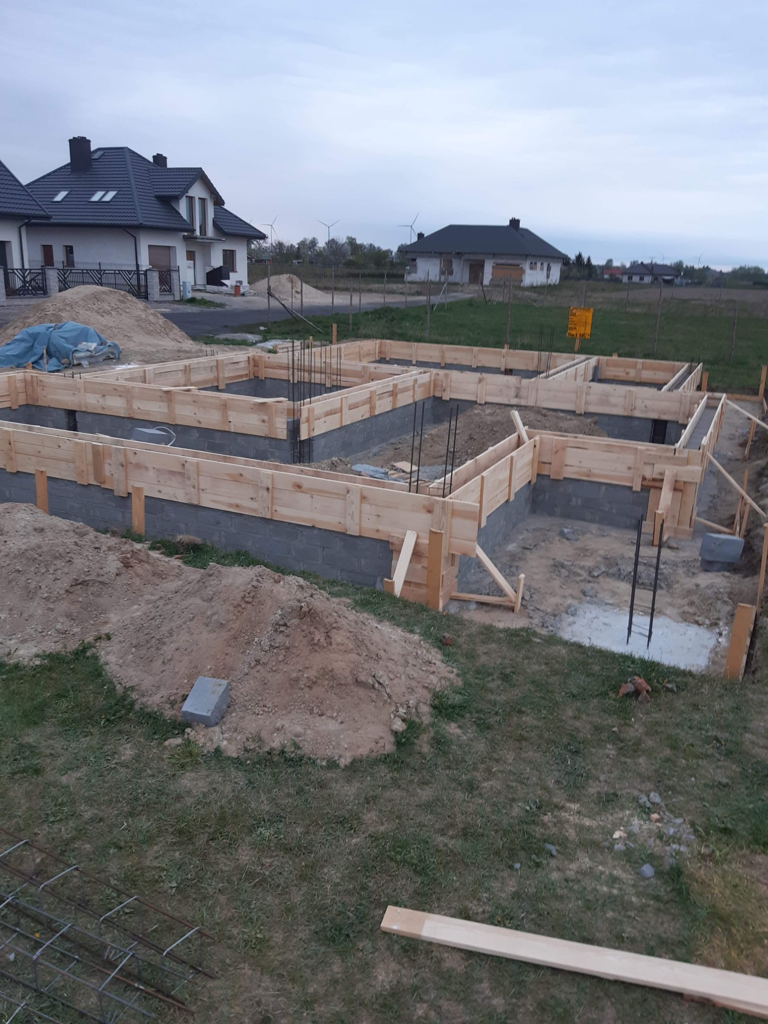 Wykopane i przygotowane fundamenty pod budowę domu jednorodzinnego, widoczne szalunki z desek, wystające pręty zbrojeniowe, w tle domy w zabudowie szeregowej i turbiny wiatrowe.