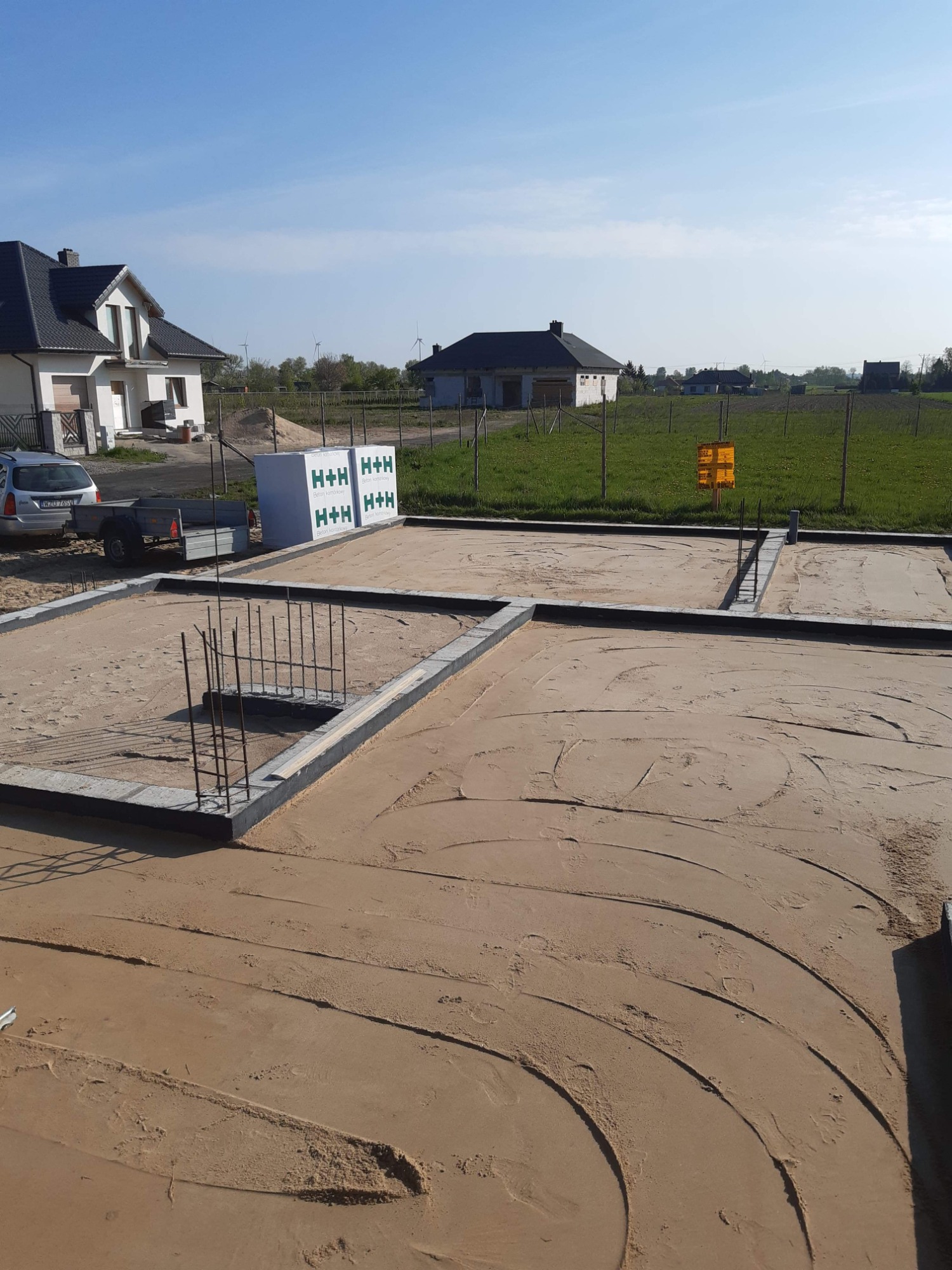 Przygotowany teren pod wylewkę fundamentów domu jednorodzinnego, widoczne zbrojenia i bloczki fundamentowe, w tle zabudowa jednorodzinna i błękitne niebo.