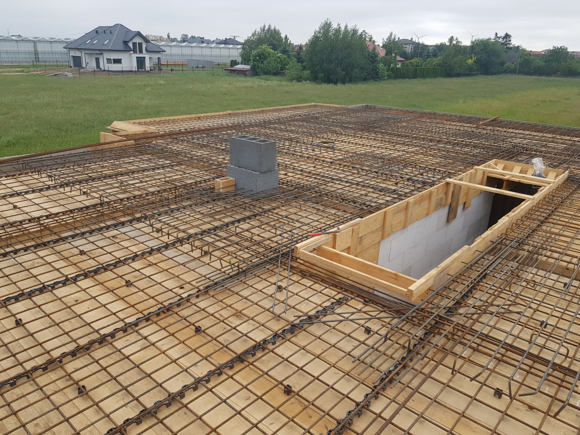 Zbrojenie płyty fundamentowej z widocznymi bloczkami betonowymi i szalunkiem otworu, na tle krajobrazu z domem i szklarniami.