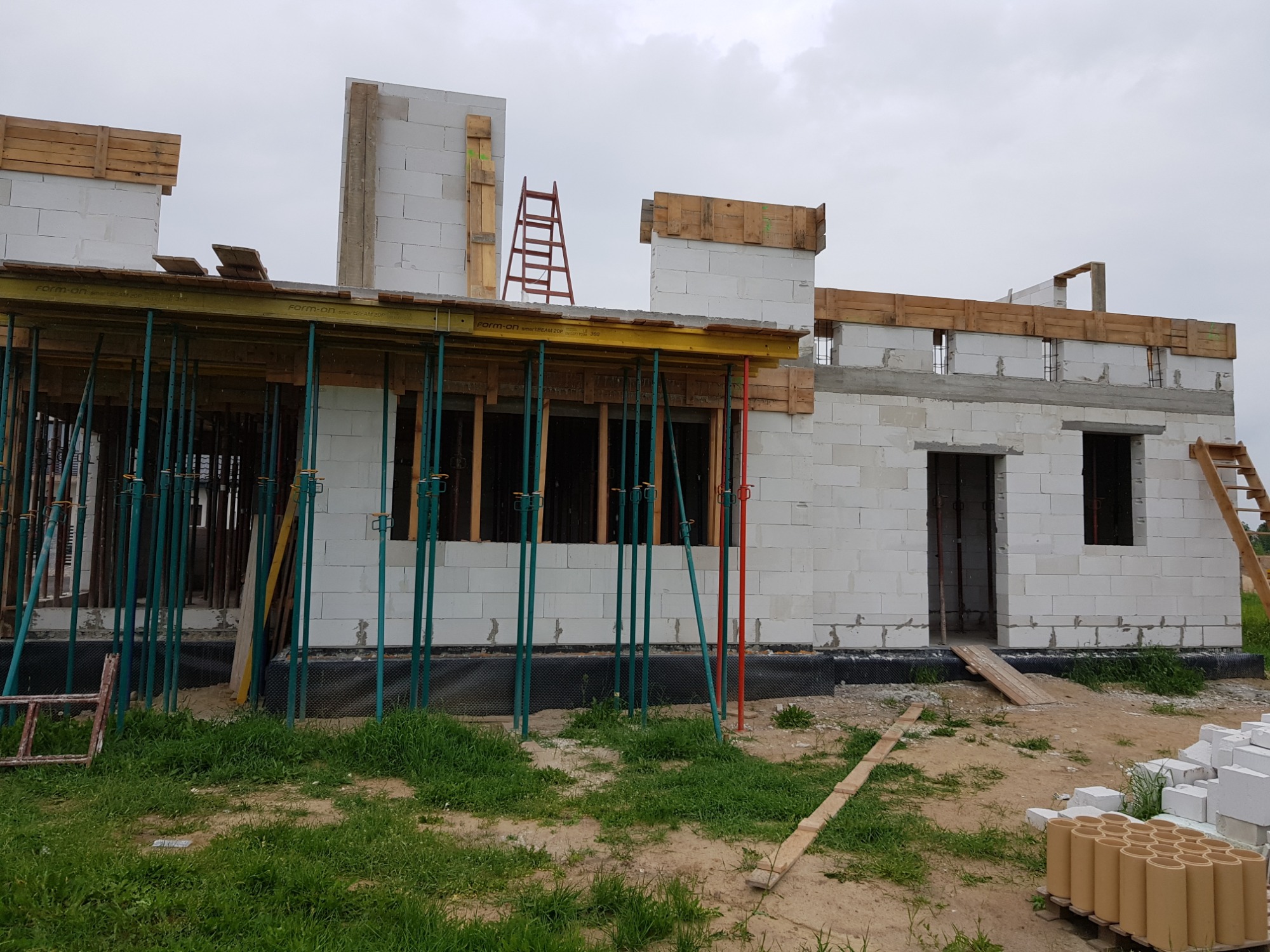 Budowa domu z białych bloczków, widoczne szalunki, podpory i drabiny, teren wokół w trakcie przygotowania.