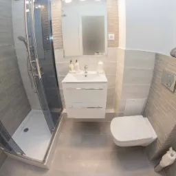 Nowoczesna łazienka w szarościach z kabiną prysznicową, umywalką z szafką i toaletą, wykończona płytkami imitującymi cegłę, z dekoracyjnymi elementami jak ręczniki i klepsydra.