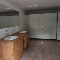 Wnętrze publicznej toalety z drewnianą szafką pod umywalkami, dużym lustrem i kabinami WC z białymi drzwiami.