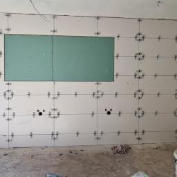 Rek-Bud - Białe płytki ścienne w trakcie układania, z krzyżykami dystansowymi i sznurkiem poziomującym, widoczny fragment ściany z płyt gipsowo-kartonowych, przygotowane otwory na instalacje, wiadro z klejem...