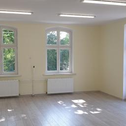 Rek-Bud - Puste, odmalowane pomieszczenie z trzema oknami, jasnymi ścianami, drewnopodobną podłogą i białymi grzejnikami.