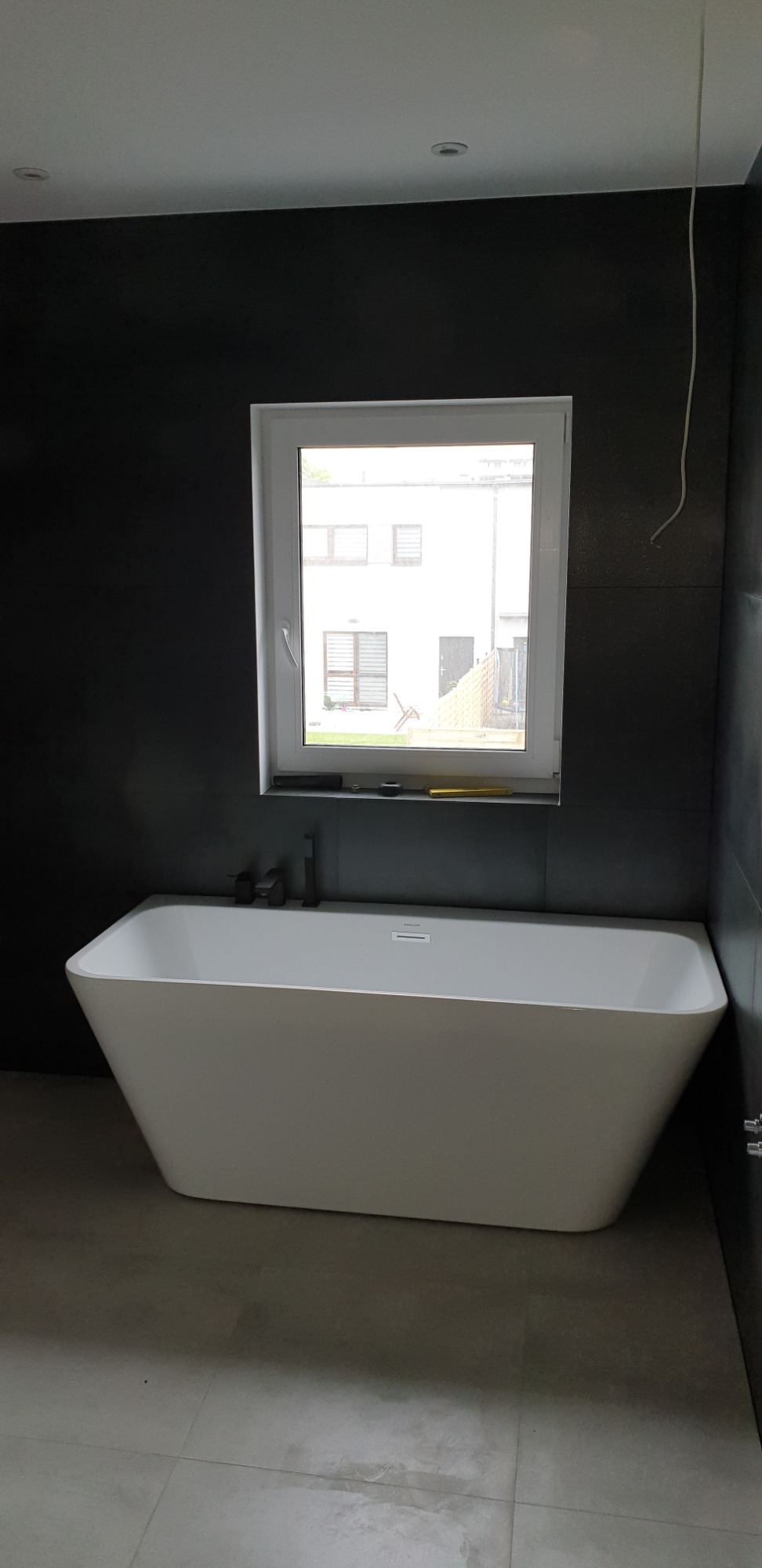 Nowoczesna wanna wolnostojąca o trapezoidalnym kształcie, z czarną armaturą, na tle ciemnych, grafitowych płytek, z oknem wychodzącym na budynek mieszkalny, widoczne narzędzia na parapecie.