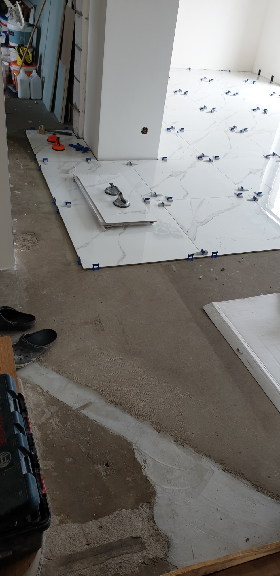 Białe, marmurkowe płytki podłogowe układane z użyciem przyssawek i niebieskich krzyżyków dystansowych, widoczne narzędzia i nieprzygotowane podłoże.