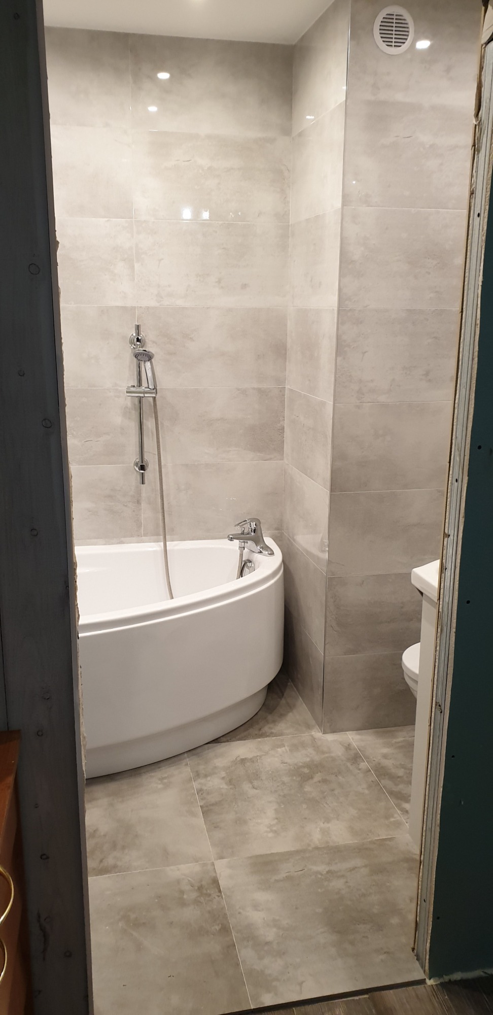 Nowoczesna łazienka z wanną narożną, wykończona płytkami w odcieniach szarości, widziana z perspektywy niedokończonego wejścia, z widocznym wentylatorem sufitowym.