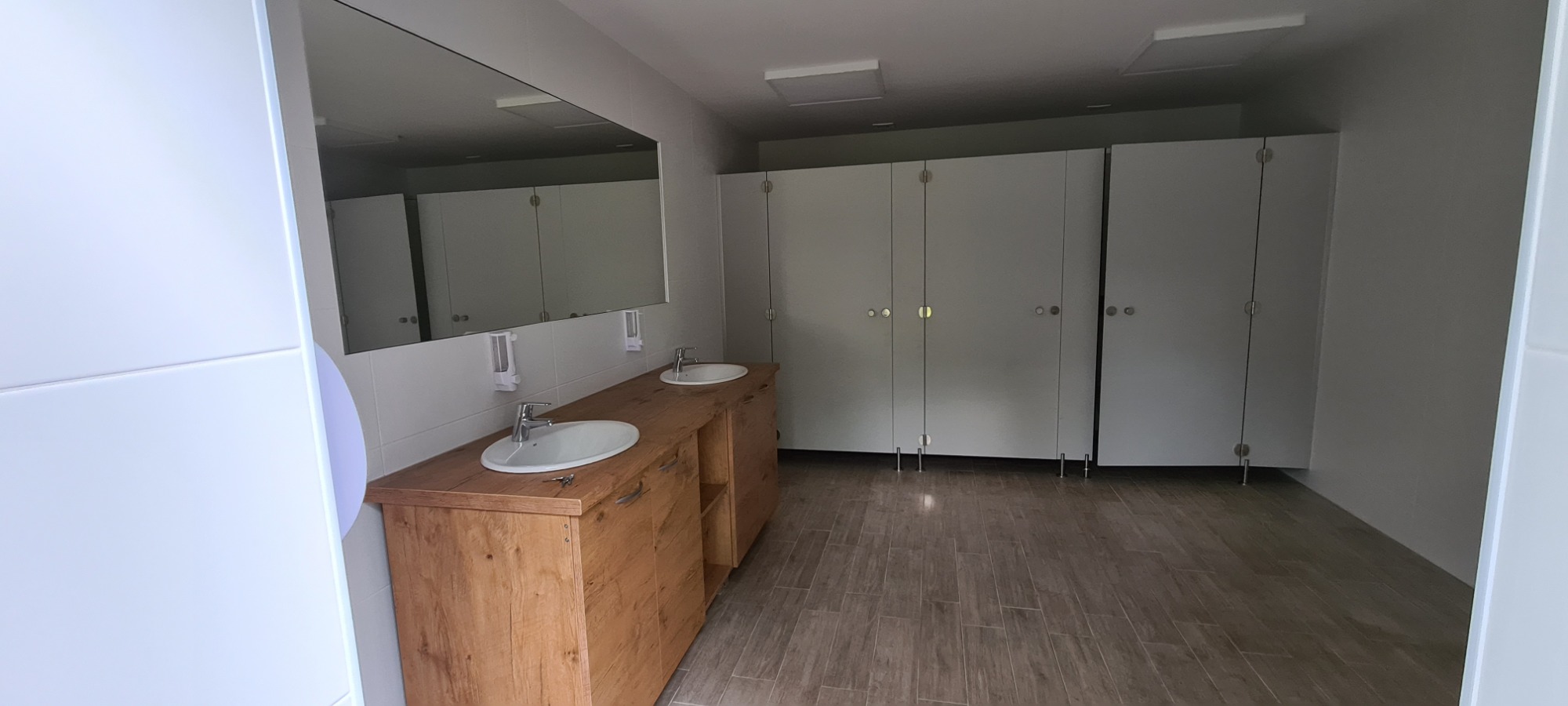 Wnętrze publicznej toalety z drewnianą szafką pod umywalkami, dużym lustrem i kabinami WC z białymi drzwiami.