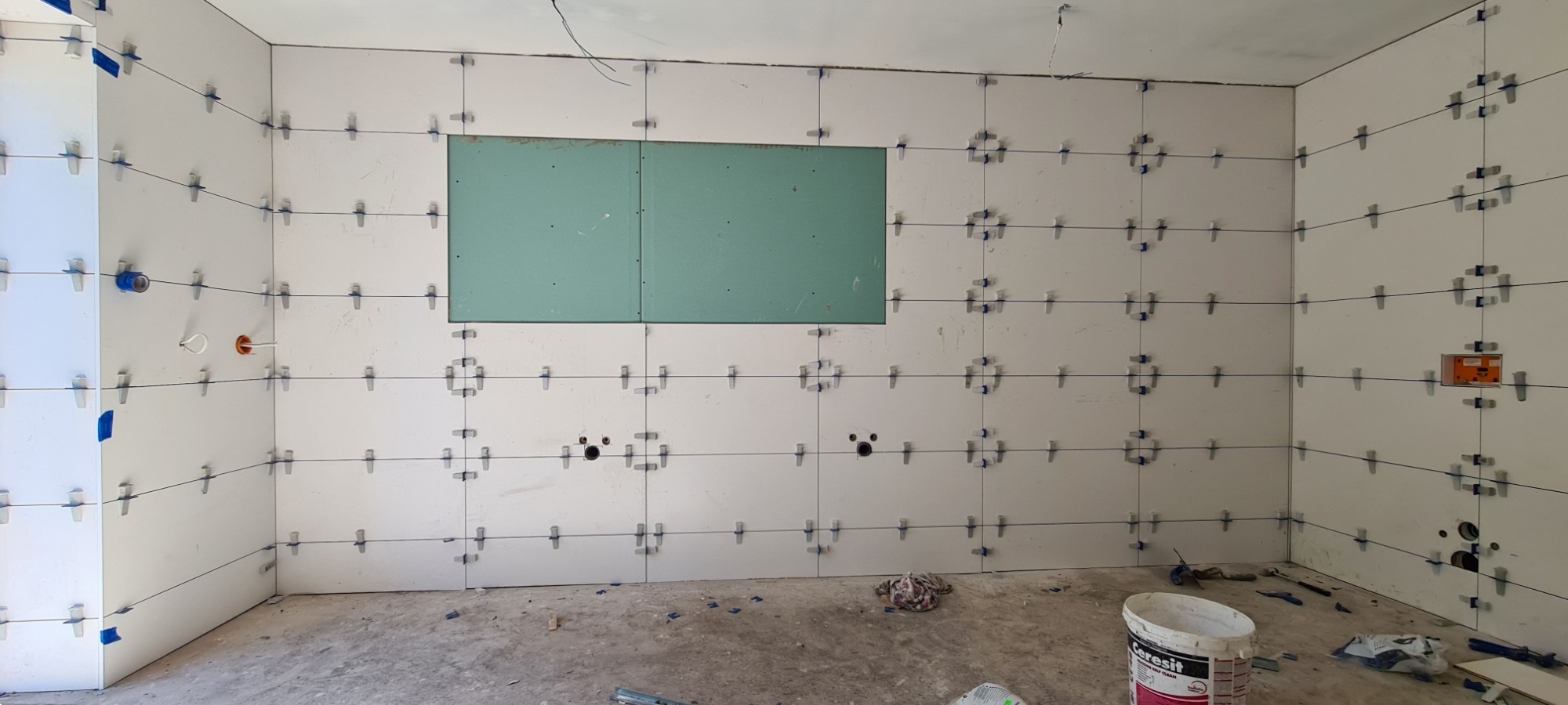 Białe płytki ścienne w trakcie układania, z krzyżykami dystansowymi i sznurkiem poziomującym, widoczny fragment ściany z płyt gipsowo-kartonowych, przygotowane otwory na instalacje, wiadro z klejem...