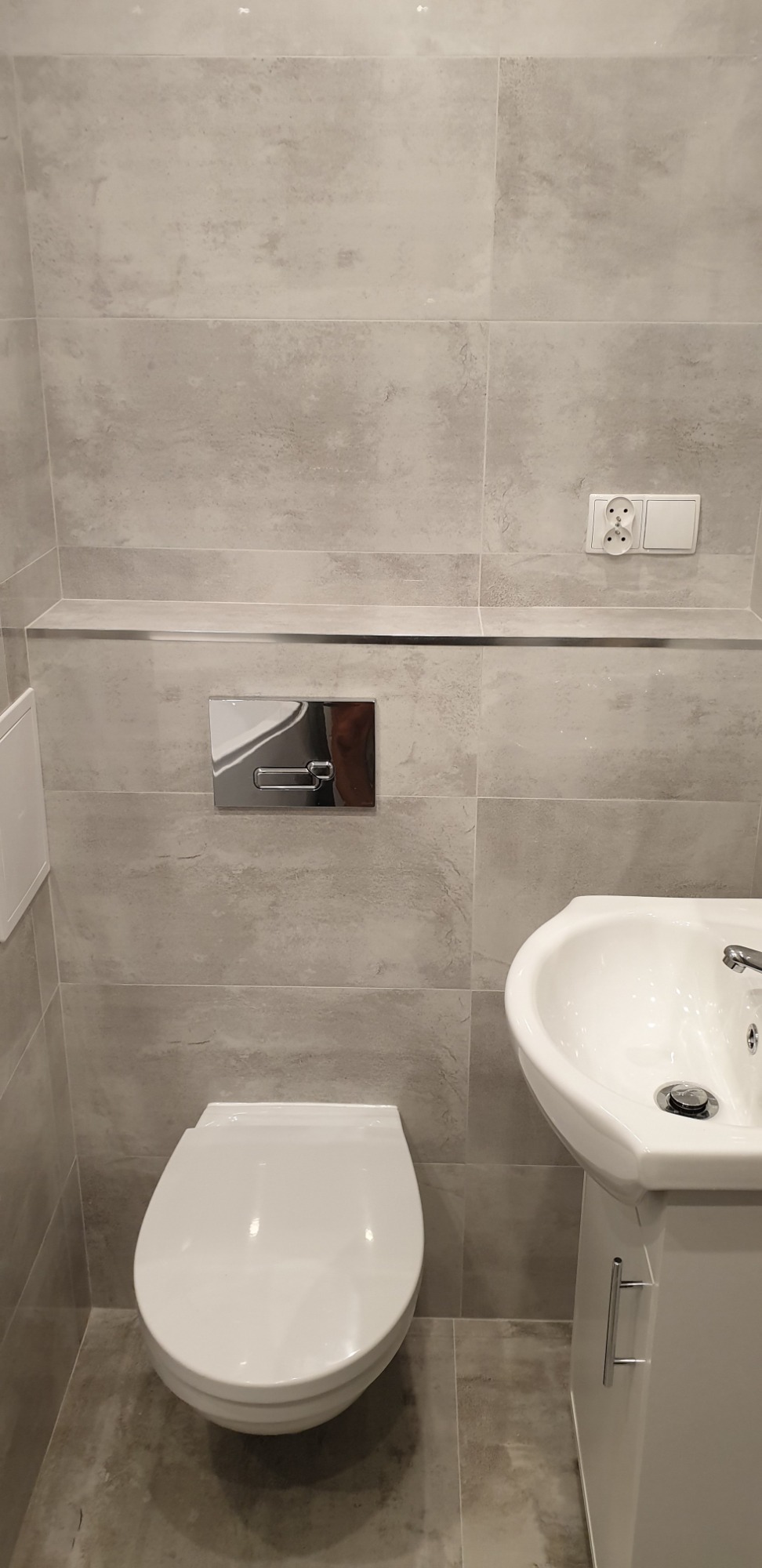 Wykończona łazienka z szarymi płytkami, wiszącą toaletą z chromowanym przyciskiem spłukiwania, białą umywalką na szafce i gniazdkiem elektrycznym.