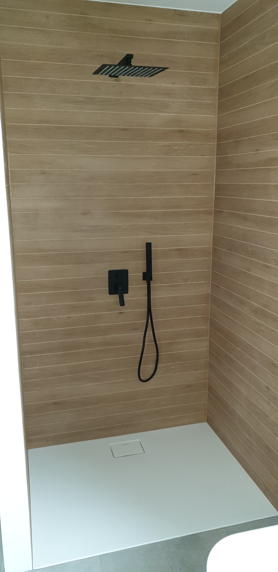 Kabina prysznicowa z odpływem liniowym, wykończona płytkami imitującymi drewno, wyposażona w czarną deszczownicę i baterię.
