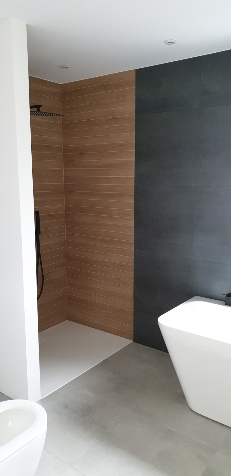 Nowoczesna łazienka z kabiną prysznicową typu walk-in, wykończona płytkami imitującymi drewno i szarą ceramiką, z białą wolnostojącą wanną i podwieszaną toaletą.