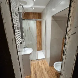 Wąskie ujęcie na wykończoną łazienkę z kabiną prysznicową, umywalką i grzejnikiem, widzianą przez surowy otwór w ścianie. Płytki imitujące drewno na podłodze i ścianach.